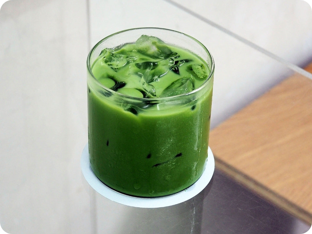 Ein Iced Matcha Latte in einem zylindrischen rundem Glas mit Eiswürfeln befüllt auf einem Glastisch stehend.