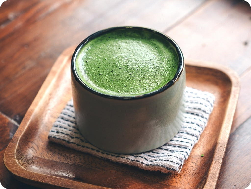 Matcha Latte mit dichtem, feinen Schaum in einer zylindrisch geformten weissen Matchaschale mit schwarzem Rand. Auf einem Tuch und einem Holztablett mit runden Ecken.