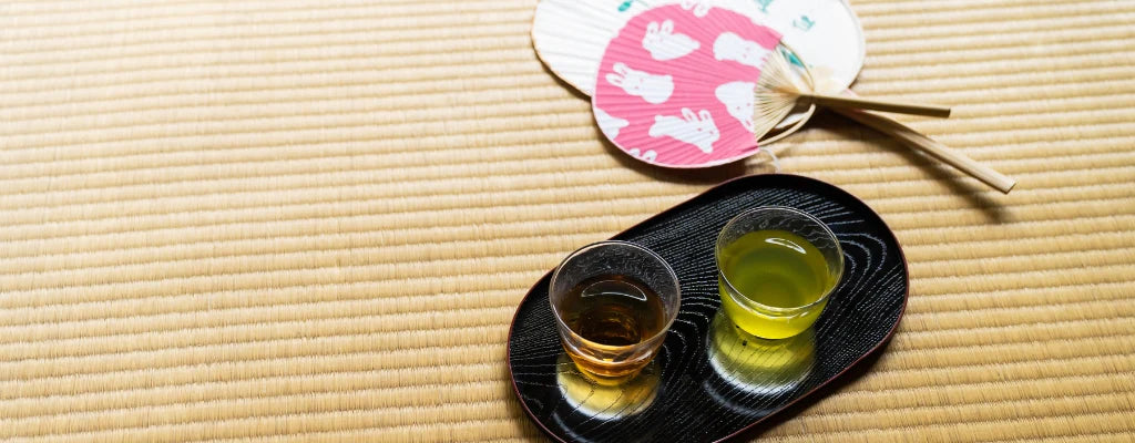 Auf einer Tatami Matte ist ein Tablett, welches mit 2 Gläsern gedeckt ist. In dem einen Glas ist ein grüner Tee und in dem anderen ein Hojicha.