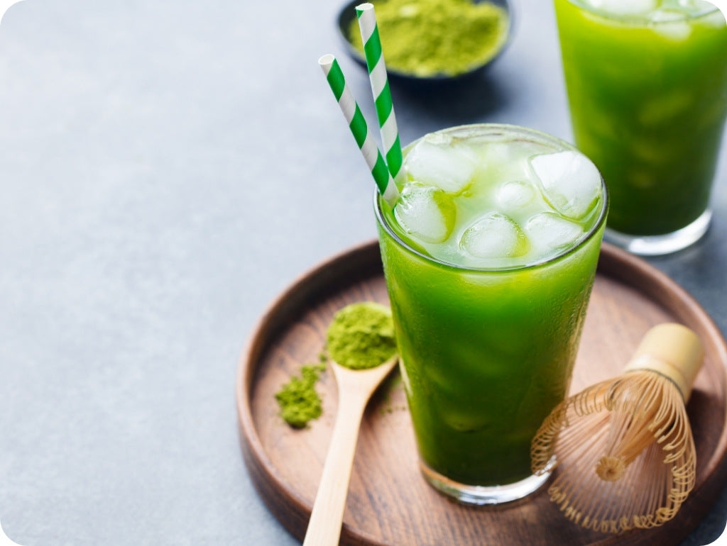 Ein Holztablett mit einem Glas, einem Matchabesen und einem Löffel darauf. Auf dem Löffel ist Matcha und in dem Glas ein Iced Matcha mit Eiswürfeln und einem Strohhalm.