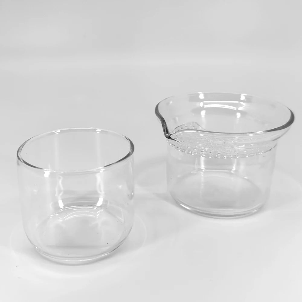 Ein Teeset bestehend aus einem kleinen zylindrischen Glas und einer Hohin Glas Teekanne welche geöffnet ist mit einem integriertem Glas Teesieb. Das Set ist vor einem weiss-gräulichen Hintergrund. Man sieht Spiegelungen im Glas und den Schatten der Produkte.