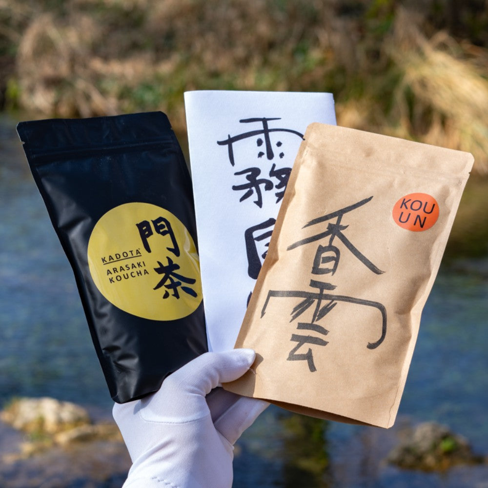 3 japanische Tee Verpackungen von einer Hand mit einem weissen Handschuh gehalten vor einem Fluss.