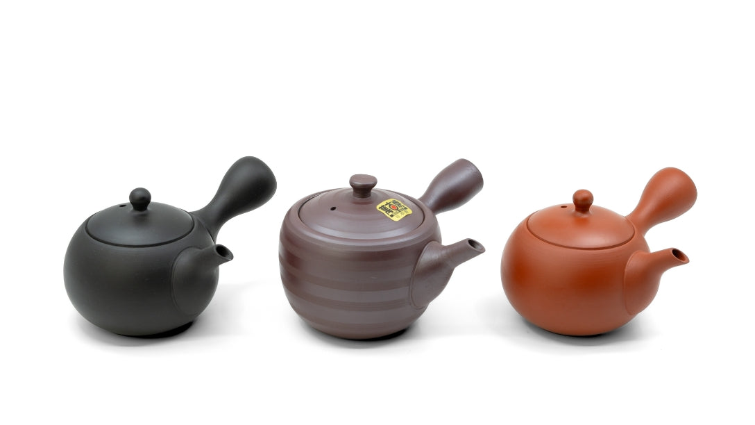 3 Kyusu Teekannen nebeneinander vor einem weissen Hintergrund freigestellt. Links eine schwarze in der Mitte eine braun lilane und rechts eine rote.