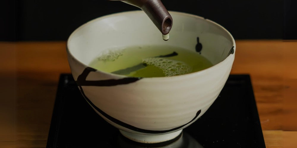 Eine weiße Teeschale mit grünem Tee auf einem schwarzen Untersetzer.