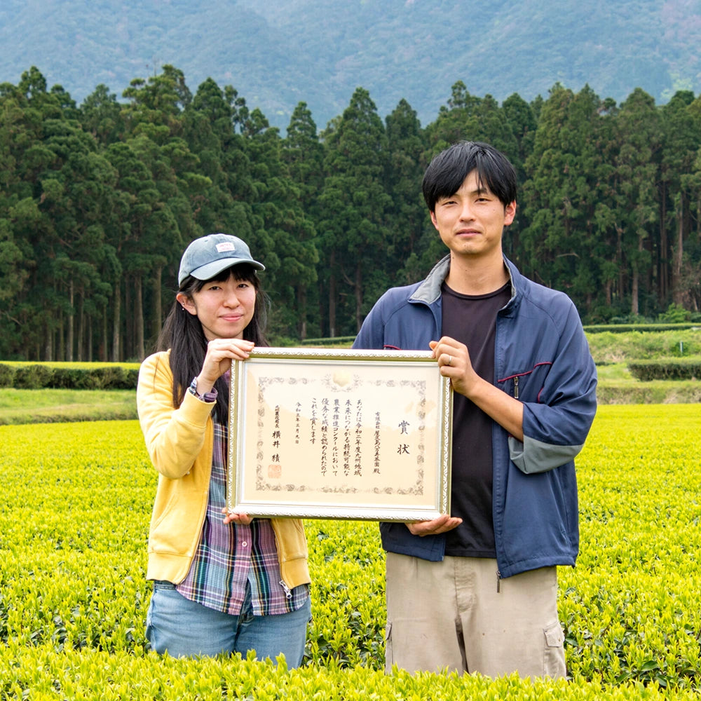 Mariko & Keita Watanabe, die vor ihrem Teefeld stehen und eine ihrer Auszeichnungen stolz präsentieren. Im Hintergrund sind Bäume und ein Berg.